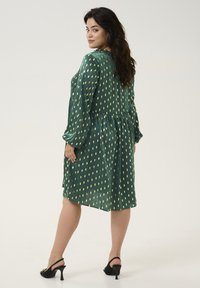 Robe verte avec motif de feuilles dorées, manches longues, taille froncée, longueur genou. Portée avec des chaussures à talons noires. Texture de tissu lisse.