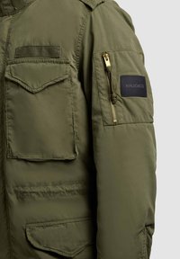 Manche de veste vert olive avec poches frontales à rabat, une poche zippée sur le bras supérieur, et un patch rectangulaire noir de la marque Khujo.