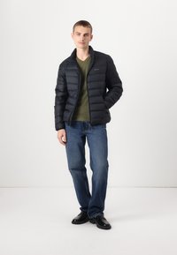 Giubbotto puffo nero con colletto rialzato, chiusura con zip e cuciture orizzontali, abbinato a una maglietta verde a scollo a V e jeans blu. Scarpe nere.