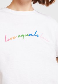 Bílý tričko s krátkým rukávem vyrobené z bavlny, které zdobí barevný text "láska se rovná lásce" v gradientních odstínech růžové, oranžové, zelené a modré.
