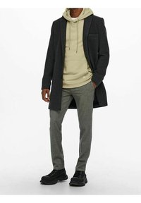 Schwarzer Wollmantel über einem hellgr�ünen Hoodie, kombiniert mit grauen gemusterten Hosen und schwarzen Sneakers. Klare Linien und ein strukturiertes Design.