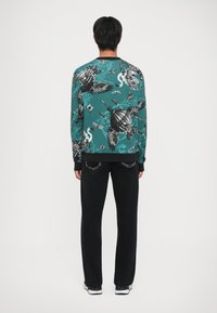 Felpa turchese con stampa grafica intricata in bianco e nero, abbinata a jeans neri e sneakers bianche. Presenta polsini e orlo a coste.