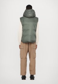 Vesta pufo gri cu o glugă mare, purtată peste un pulover deschis, asociată cu pantaloni cargo bej și încălțăminte neagră. Design simplu, utilitar.