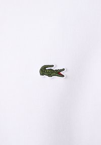 Tissu blanc à finition texturée arborant un logo crocodile vert brodé avec des accents rouges, placé sur la partie gauche de la poitrine.