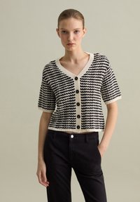 Cardigan corto a righe nero e crema con scollo a V e grandi bottoni neri, caratterizzato da un design cropped e un tessuto a maglia texturizzato.