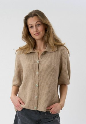 Pikkade juustega naine, kellel on seljas beež lühikeste varrukatega nööbitud kardigan ja tumedad püksid, käed taskutes, lihtsa hele taustaga.