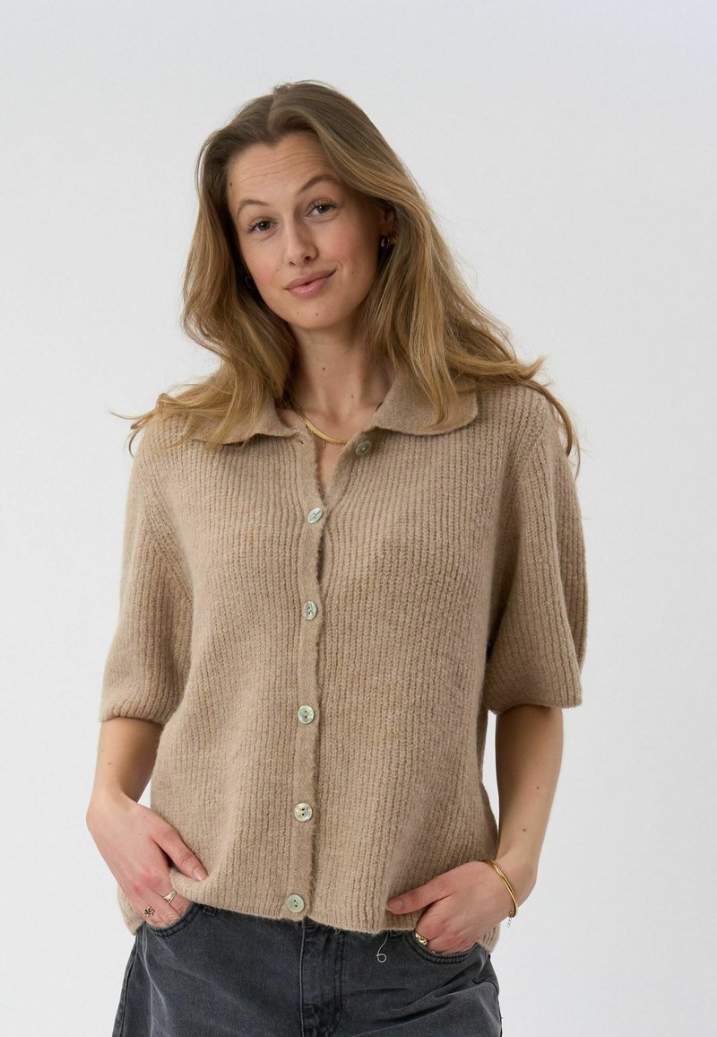 Femme aux longs cheveux portant un cardigan beige à manches courtes boutonné et un pantalon sombre, les mains dans les poches, devant un fond clair uni.