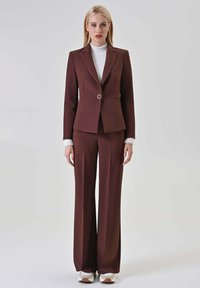 Completo su misura color marrone con blazer aderente e pantaloni a gamba larga, caratterizzato da un bottone rotondo argentato. Dolcevita bianco testurizzato sotto.