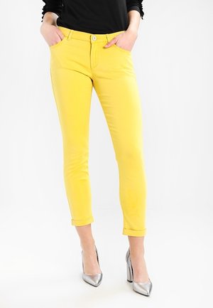 Kobieta ubrana w dopasowane, żółte, krótkie spodnie oraz srebrne buty na wysokim obcasie, z rękami w kieszeniach, stojąca na białym tle.