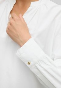 Blusa bianca con collo alto, dettagli arricciati sulle spalle e polsini con bottoni caratterizzati da un accento dorato. Tessuto dalla texture liscia.