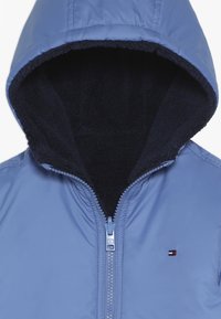 Jasnoniebieska kurtka z kapturem na zamek błyskawiczny, z miękką czarną podszewką z polaru wewnątrz kaptura. Posiada gładką powierzchnię na zewnątrz oraz małe logo marki.