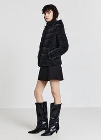 Schwarze Steppjacke mit hohem Kragen, gestepptem Design, Frontreißverschluss und zwei Seitentaschen; kombiniert mit einem schwarzen Minirock und glänzenden kniehohen Stiefeln.