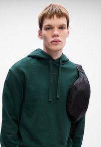 Czarna nylonowa torba na ramię o zaokrąglonym kształcie, noszona na ciemnozielonym hoodie z kapturem, który ma sznurki ściągające i logo na klatce piersiowej.