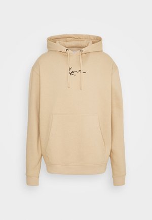 Homme portant une veste bomber verte sur un sweat à capuche beige, un pantalon noir et des chaussures de course blanches, debout les mains dans les poches.