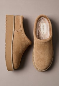 I clogs in suede color tan presentano una morbida fodera interna, una suola piatta con scanalature textured e una cucitura decorativa sul lato.