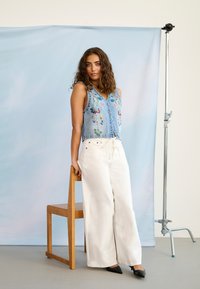 Femme aux cheveux bouclés portant un gilet en denim brodé de fleurs et un pantalon blanc à jambes larges, appuyée contre une chaise en bois dans un studio.