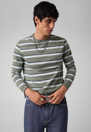 Jonge man die een groen-wit gestreept shirt met lange mouwen en blauwe jeans draagt, zijn riem verstelend, een ketting draagt en ringen.