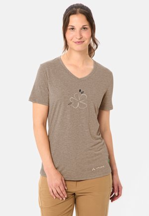 Frau trägt ein braunes V-Ausschnitt-T-Shirt mit einfachem Blumendesign und beigefarbenen Hosen, stehend vor einem einfarbigen hellen Hintergrund.