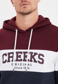 Felpa con cappuccio a blocchi di colore marrone e blu navy con dettagli bianchi, caratterizzata da una grafica logo frontale e lacci, realizzata in tessuto morbido.