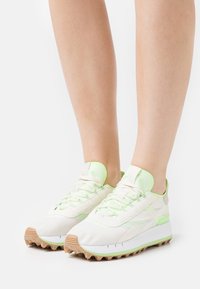 Baskets de sport blanches avec des accents vert clair, dessus en maille texturée, semelle en caoutchouc avec des picots et lacets plats. Idéales pour les activités en plein air.