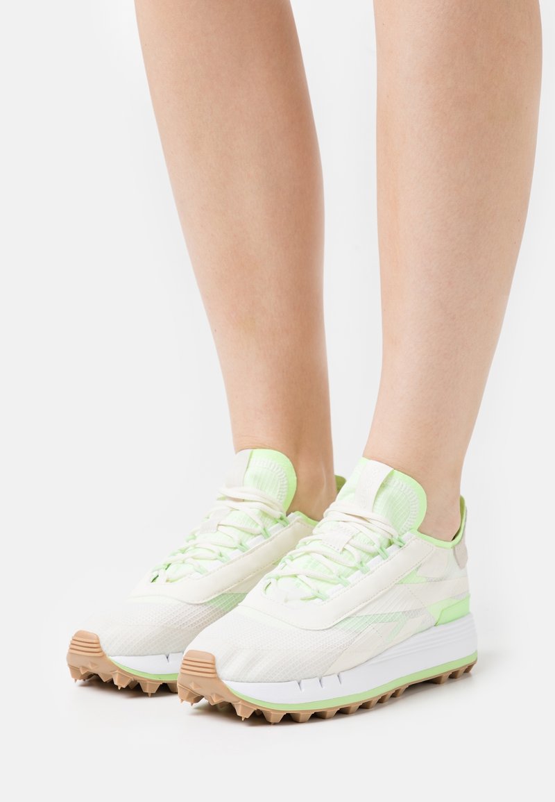 Baskets de sport blanches avec des accents vert clair, dessus en maille texturée, semelle en caoutchouc avec des picots et lacets plats. Idéales pour les activités en plein air.