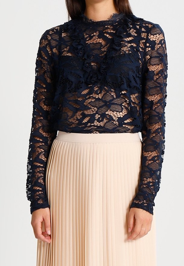 Haut transparent en dentelle bleu marine à manches longues avec des motifs floraux, associé à une jupe plissée beige taille haute.