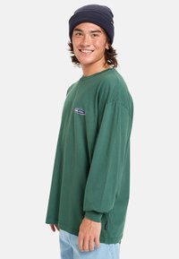 Langarm groene shirt van katoen met een ronde halslijn en bedrukte logo. Gecombineerd met lichtblauwe jeans, draagt het model een marineblauwe beanie.