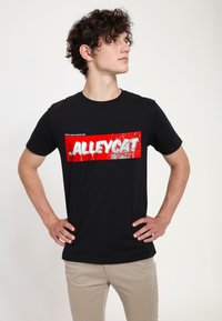 Černé bavlněné tričko s výraznou červenou obdélníkovou grafikou, na které je napsáno "ALLEYCAT" bílým písmem, na jednoduchém pozadí.