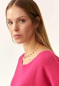 Maglione a maglia fucsia con scollatura ampia e texture a coste, abbinato a una delicata collana con piccole perline colorate.