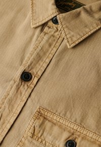 Chemise beige à boutons avec un tissu texturé, présentant des rayures verticales, une poche poitrine et des boutons noirs. Étiquette visible au niveau du col.