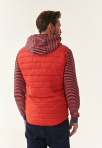 Rød polstret vest med vandret syninger og en stribet hætte i marineblå og rød. Bruges over en langærmet skjorte med lignende striber.