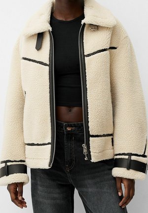 Personne à la peau foncée portant une veste en polaire sherpa crème avec des bordures et des boucles noires, un crop top noir et un jean foncé.