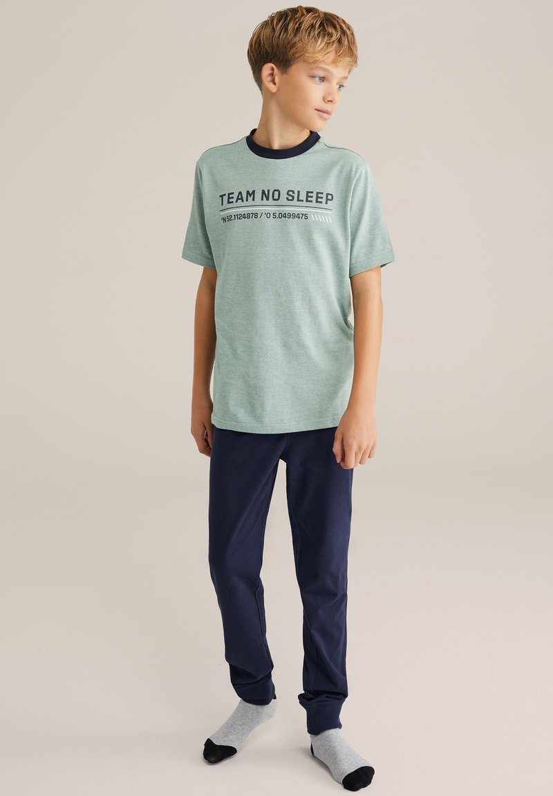 Svetlozelené tričko s nápisom "TEAM NO SLEEP" a tmavomodrým golierom, doplnené o tmavomodré nohavice a šedé ponožky s čiernymi prstami.