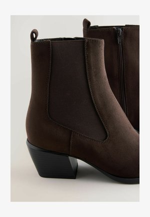 Ruskohuiset ruskeat nahkasaappaat, joissa on kaksinkertaiset elastiset sivupaneelit, vetoketjukiinnitys ja matala lohkopohja. Suuri ja sileä pinta, jossa on ompelu yksityiskohtia.