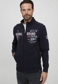 Felpa con zip blu marina in misto cotone, con grafiche testuali bianche e rosse, tasche laterali e dettagli di cucitura a contrasto.