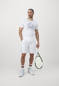 Man i vit Lacoste-t-shirt, vita shorts och randiga strumpor, hållande ett tennisracket, stående mot en enfärgad bakgrund.
