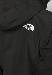 Czarna wodoodporna kurtka z kapturem, z białym logo po lewej górnej stronie. Tkanina ma gładką, matową teksturę.