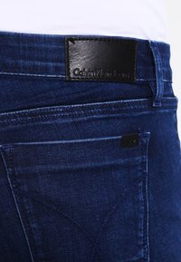 Jeans de mezclilla azul oscuro con una textura suave, que presentan una etiqueta de cinturón de cuero negro y un sutil detalle de costura en el bolsillo trasero.