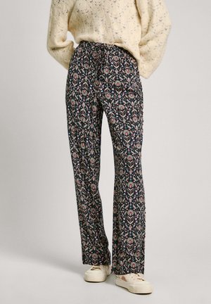 Personne portant un pantalon ample à motif floral foncé, un pull en maille crème et des baskets blanches décontractées, debout sur un fond clair.