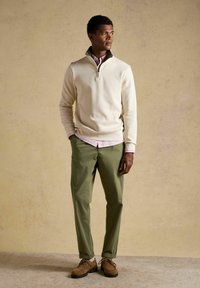 Homme portant un pull beige à fermeture éclair courte, une chemise à rayures roses, un pantalon vert olive et des chaussures marron, debout les mains dans les poches, regardant vers la droite.