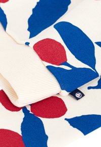 Strickpullover mit einer cremefarbenen Basis, verziert mit auffälligen blauen Blättern und roten runden Formen. Gerippte Bündchen und ein kleines Markenetikett an der Seite.