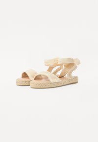 Sandales espadrilles avec larges sangles en tissu beige clair, semelle en jute tissée et design doux et courbé. Présente un style à enfiler.