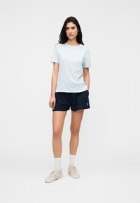 Ανοιχτό μπλε βαμβακερό μπλουζάκι με κοντά μανίκια, συνδυασμένο με σκούρα μπλε σορτς. Το μοντέλο φοράει μπεζ παπούτσια και λευκές κάλτσες, στέκεται μπροστά σε ένα απλό φόντο.