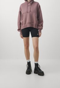 Persona vistiendo una sudadera con capucha color malva, pantalones cortos de ciclismo negros de Nike, calcetines blancos y botas negras con cordones, de pie contra un fondo liso de color gris claro.