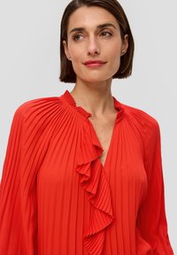 Rode, geplooide blouse met een rimpelende halslijn en lange mouwen. De stof lijkt soepel met een lichte glans en heeft verticale plooien overal.