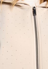 Sweat à capuche beige zippé, en tissu lisse agrémenté de clous métalliques dispersés et d'une fermeture éclair argentée. Comprend une capuche pour une protection supplémentaire.