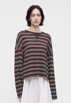 Jeune personne aux cheveux courts et foncés portant une chemise noire ample à manches longues avec des rayures blanches et rouges et une jupe blanche.