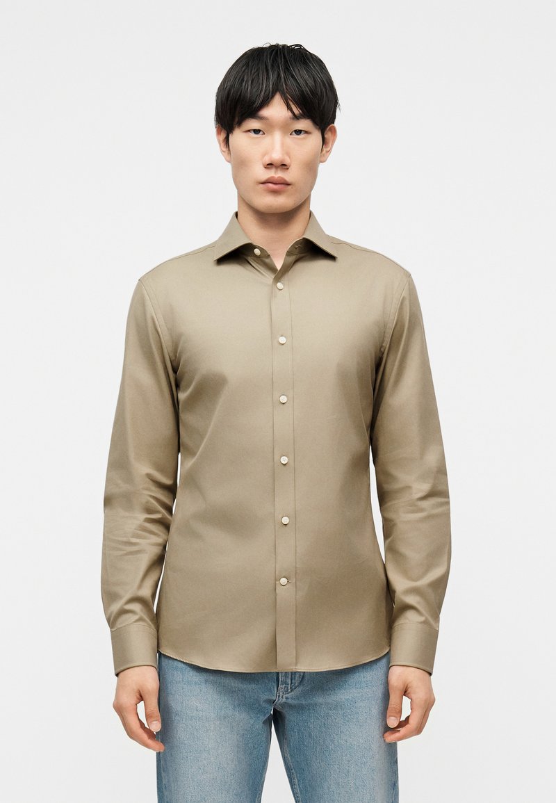 Νέος άνδρας που φοράει μπεζ μακρυμάνικο πουκάμισο με κουμπιά και ανοιχτό μπλε τζιν, στέκεται μπροστά σε απλό λευκό φόντο.