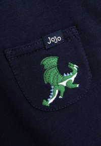 Jūras spēku kokvilnas krekls ar zaļu un baltu izšūtu pūķa motīvu uz kabatas, virs kabatas ir "Jojo" etiķete.