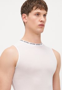 Haut sans manches en maille blanche avec un motif texturé. Présente un col noir et blanc de marque. Design ajusté avec une coupe athlétique.